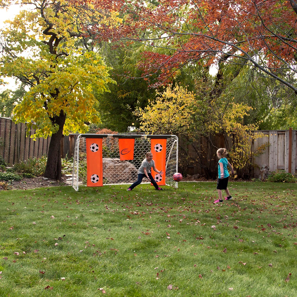 One girl kicks the ball while another girl acts as goalie, guarding the soccer goal.